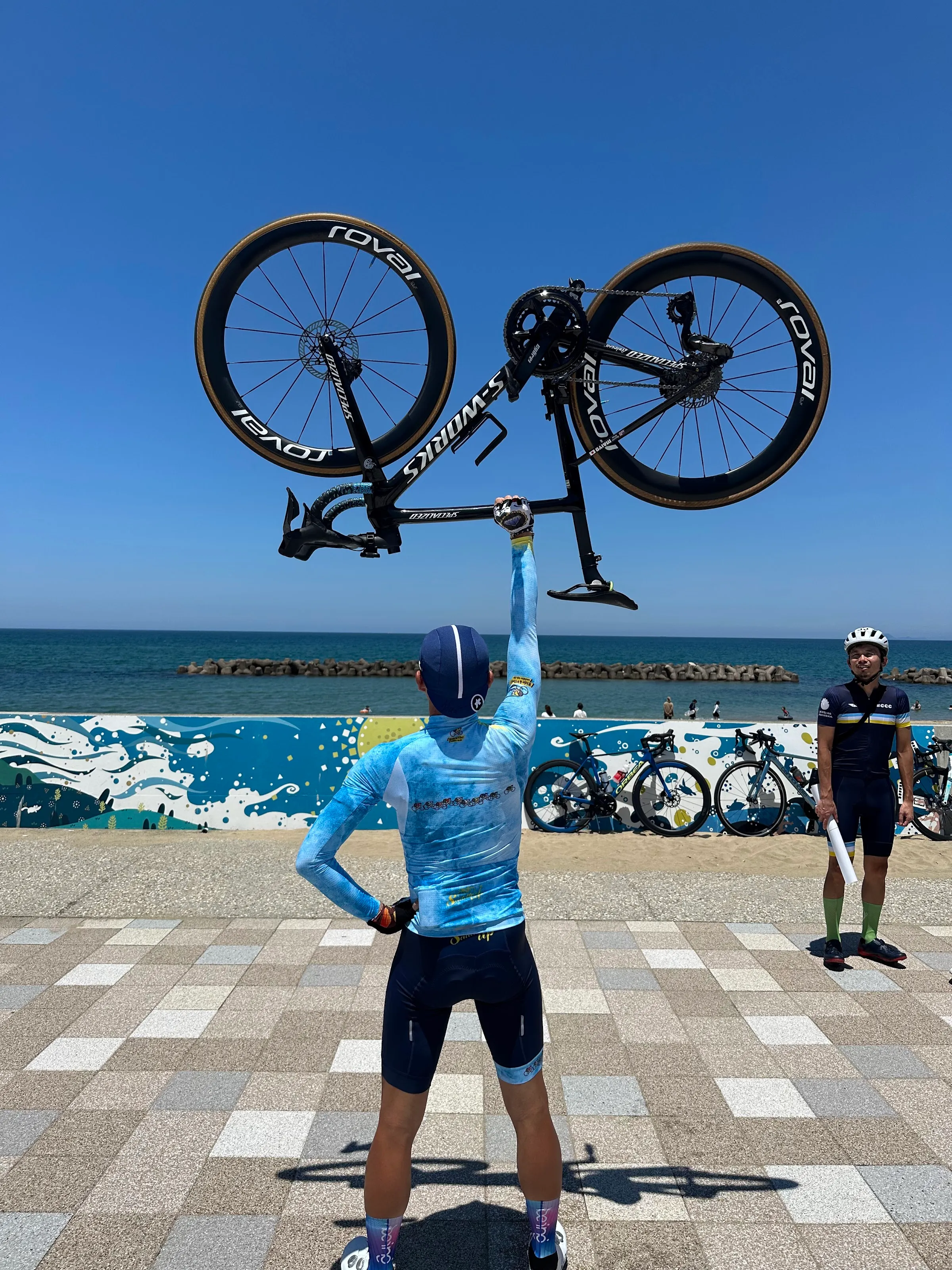 青空と海を背に、自転車を頭上に掲げるメンバー
